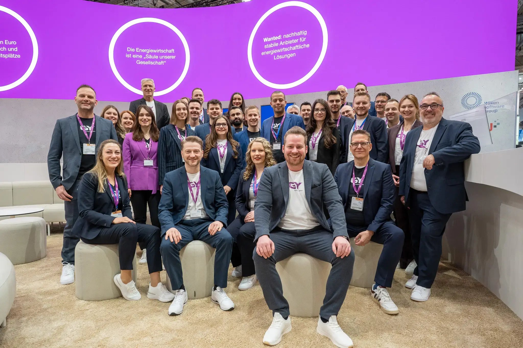 Ein Gruppenbild von einem jungen und dynamischen Messeteam. Die ersten zwei Reihen sitzen locker auf Hockern und der Rest der Personen stehen hintereinander. Im Hintergrund ist eine lila LED-Wand mit Botschaften zu sehen. Alle Personen tragen auch lila Lanyards um den Hals.